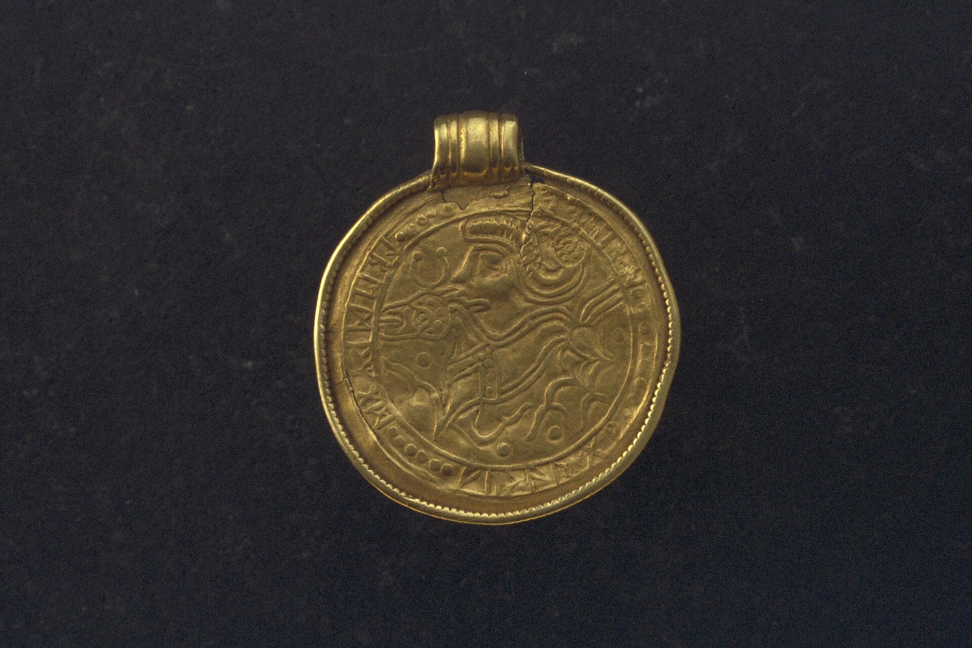 A round gold bracteate from Grumpan, Sweden, with a stylized human head above an animal in the center and runes around the rim
